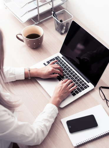 Businesswoman typing on laptop with smartphone and coffee on desk, representing professional data entry services.